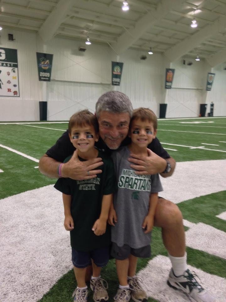 Coach Mannie kneeling on the indoor practice field with two young boys in Michigan State shirts